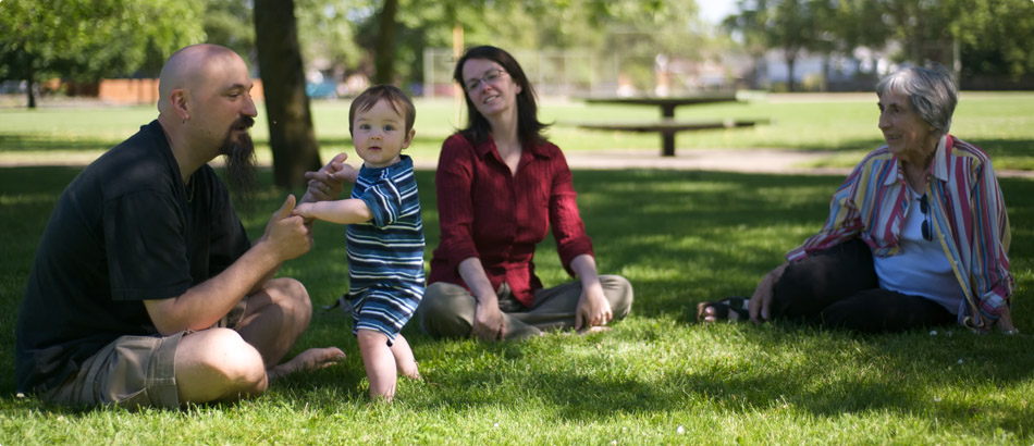 Randall, River, Angie, and Shirley on the grass in a park.  River is about a year old, standing and holding Randall's hand for support, looking right at the camera.  Everyone else is sitting and looking at River and smiling.